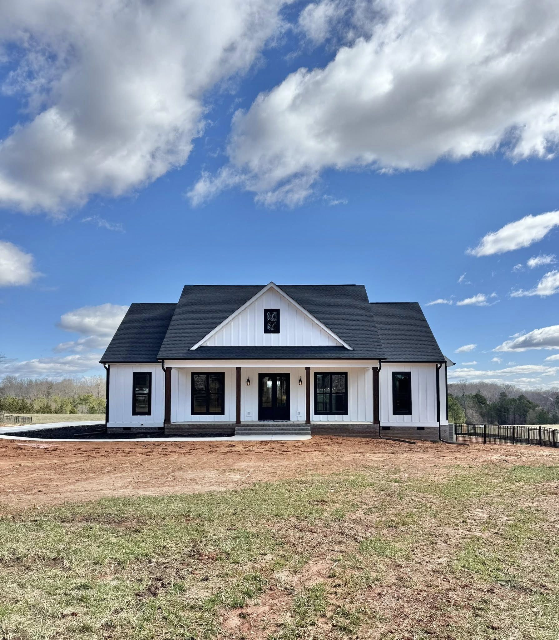 White, modern farmhouse with black roof sits under a blue sky with clouds. Red dirt and green grass cover the foreground, exuding a calm, serene atmosphere.