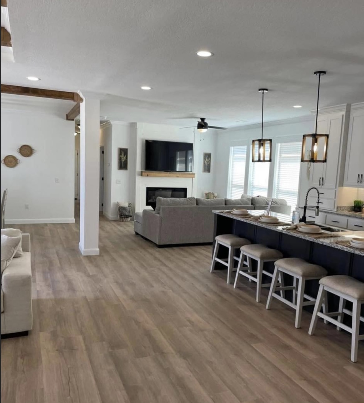 Modern open-concept living space with light wood floors, kitchen island with stools, beige sofa, fireplace, wall-mounted TV, and large windows for natural light.