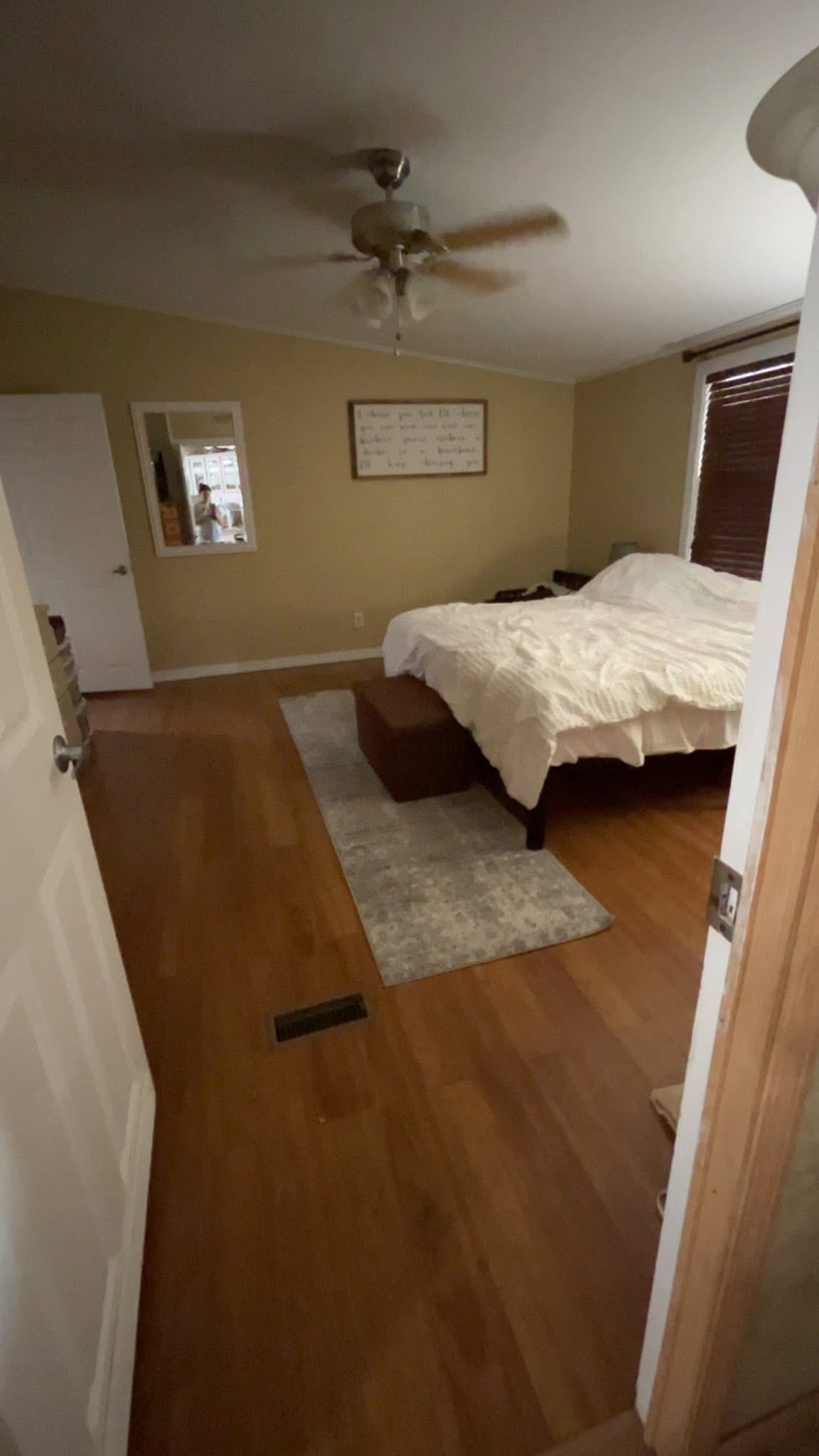 A cozy bedroom with hardwood floors and beige walls. A neatly made bed sits on a gray rug, with a ceiling fan above. A mirror and framed art adorn the walls.
