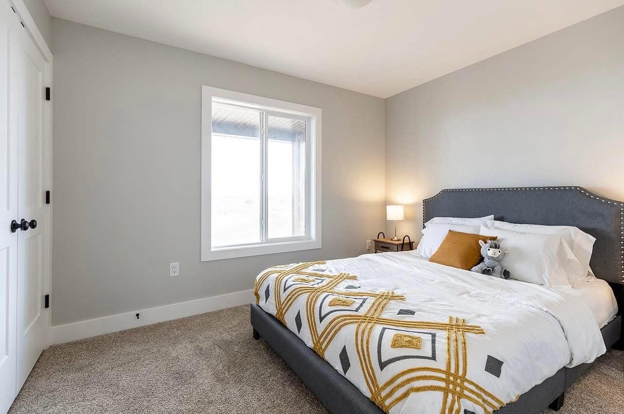 A cozy bedroom with a large, gray upholstered bed featuring a white and yellow geometric duvet. A teddy bear and pillows add a welcoming touch. Soft lighting from a bedside lamp and natural light from a window enhance the serene atmosphere.