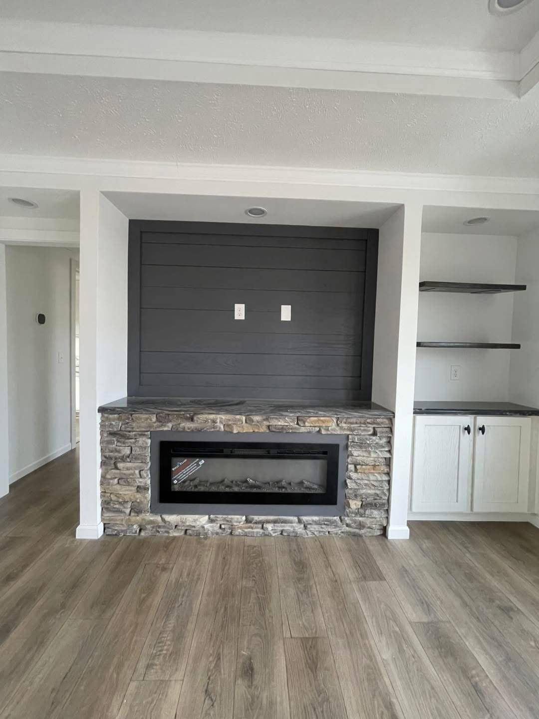 Modern living room with a stone fireplace, gray shiplap wall accent, recessed lighting, wooden shelving, and light wood floors. Cozy and stylish atmosphere.