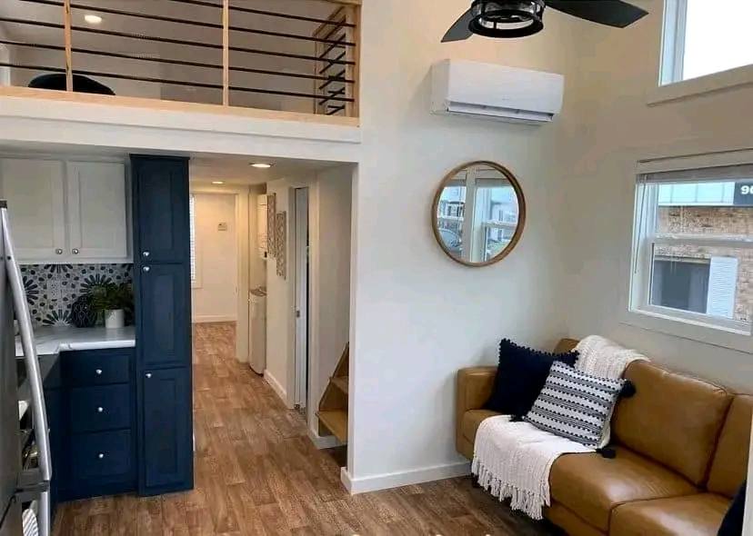 Cozy living space with a tan leather couch adorned with cushions and a throw. A loft with wooden railings overlooks the area. A navy and white kitchen contrasts with wooden floors. A round mirror and windows add brightness. Warm and inviting ambiance.