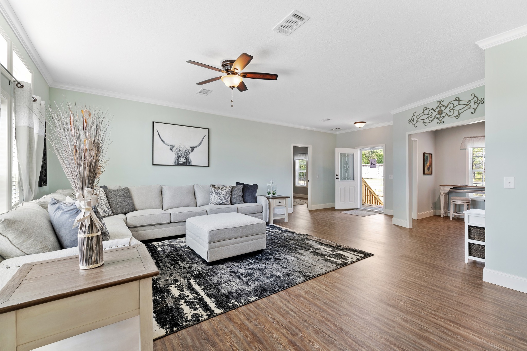 Spacious living room with a gray sectional sofa and ottoman on a dark patterned rug. Neutral tones, hardwood floors, decorative art on walls.