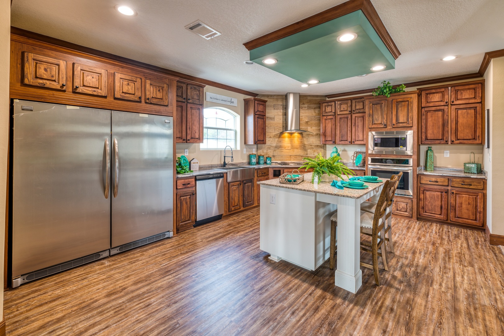 Spacious kitchen with wooden cabinets, stainless steel appliances, and an island with teal tableware. Warm lighting and plants create a cozy ambiance.