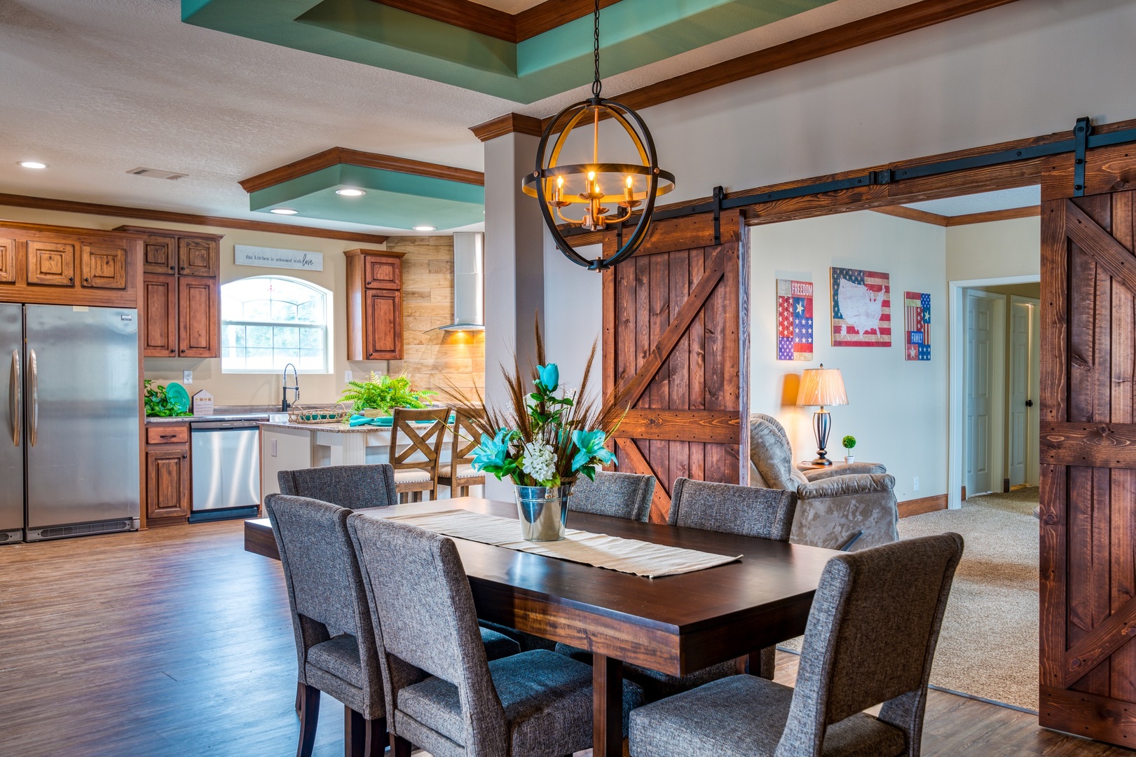 Open-concept dining and kitchen area with rustic wood elements. Cozy gray chairs surround a wooden table topped with a flower vase. Warm lighting and barn doors add charm.