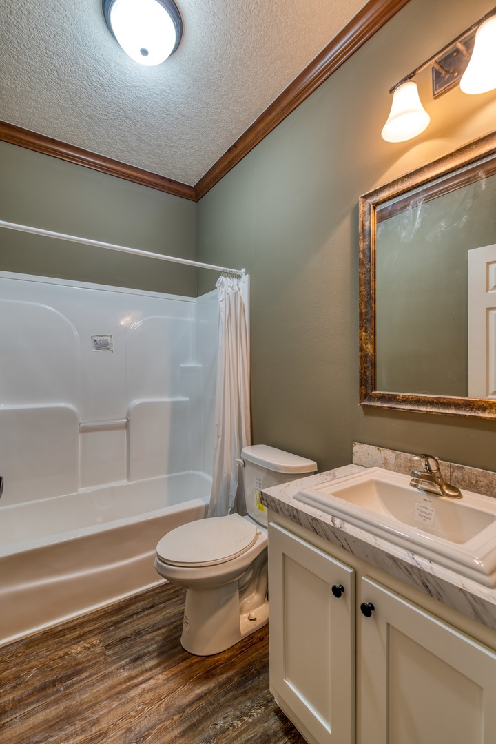 Compact bathroom with green walls and wood trim. Includes a white bathtub with curtain, toilet, and marble sink under a large mirror. Warm lighting.