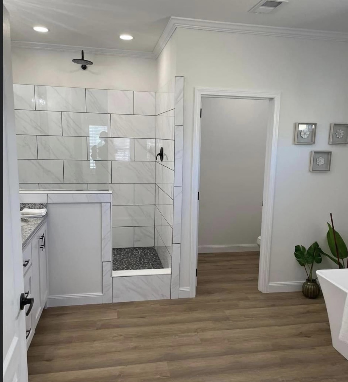 Modern bathroom with a walk-in shower featuring white tiles and black fixtures. Wood floors, a potted plant, and wall art create a serene ambiance.