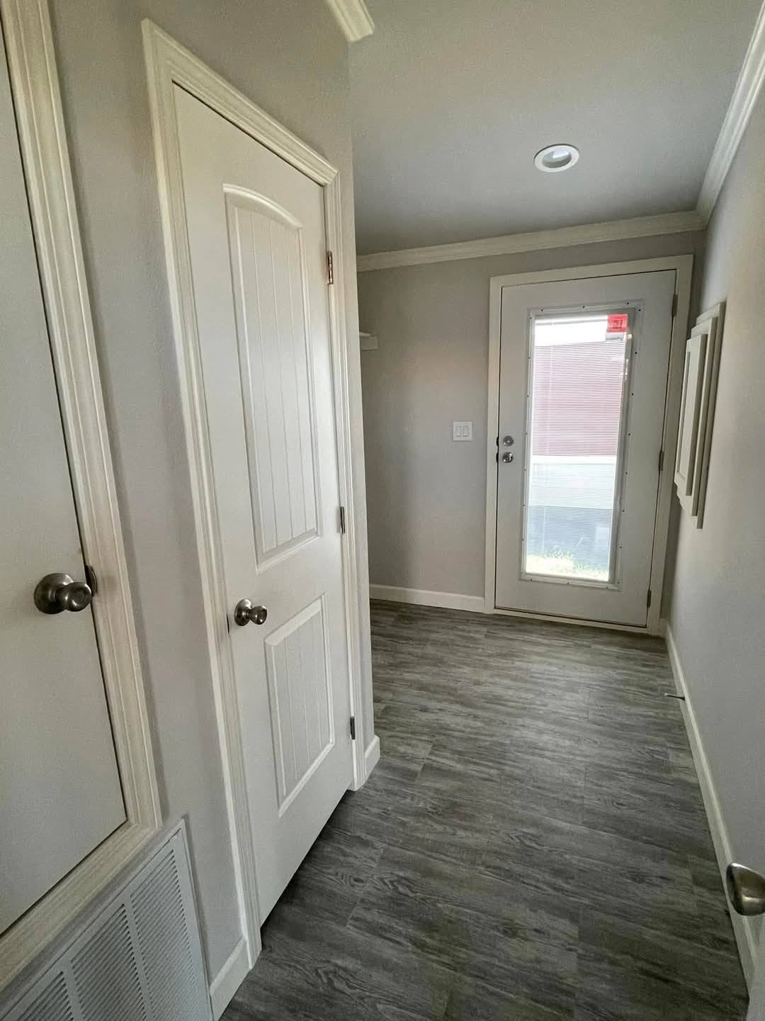 A narrow hallway with light gray walls and wood-patterned flooring. It features a closed white door on the left, a glass door at the end, and a calm ambiance.