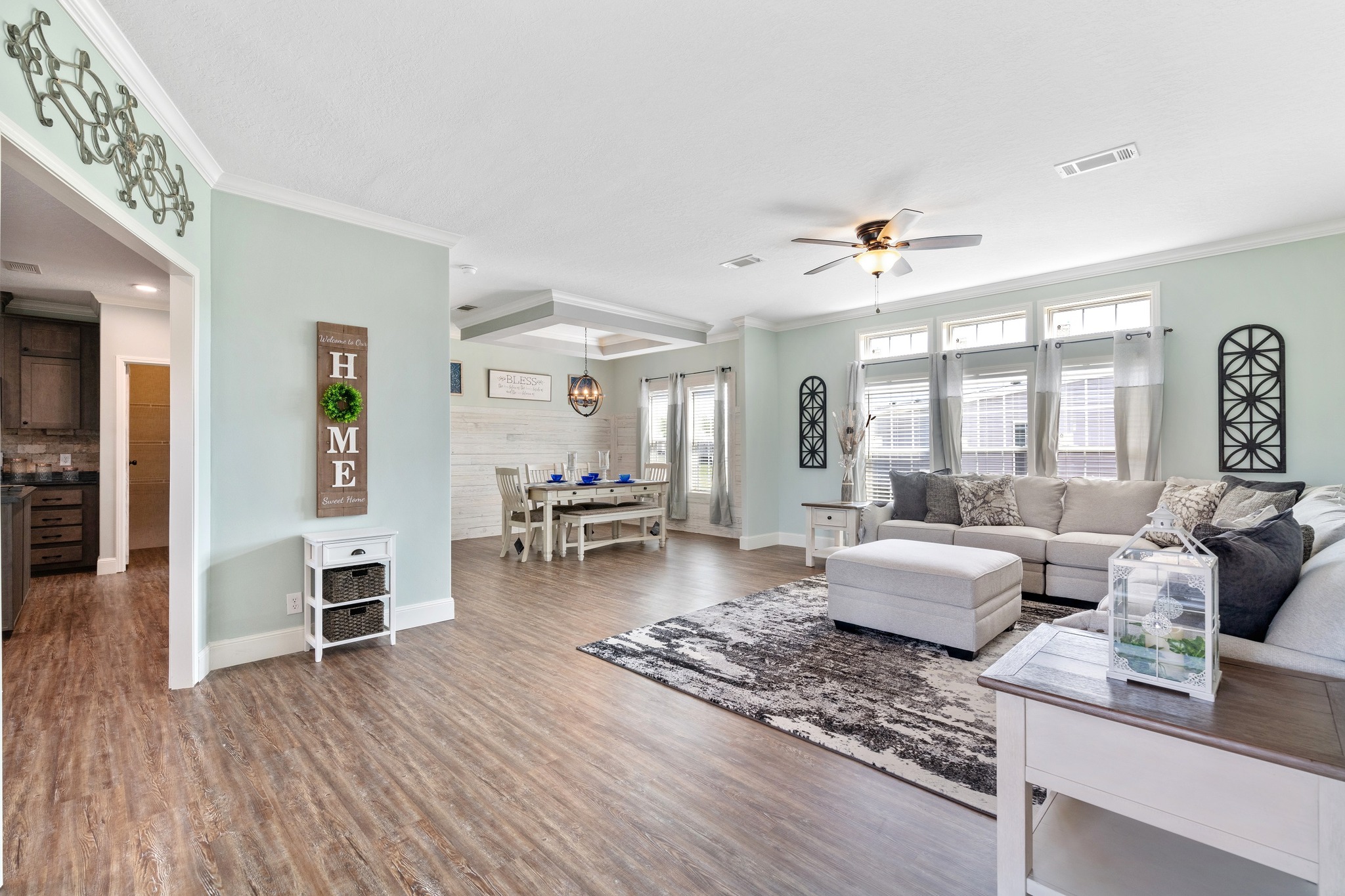 Spacious, airy living room with light teal walls, wood flooring, a gray sectional sofa, dining area with white table, and large windows. Cozy, welcoming vibe.