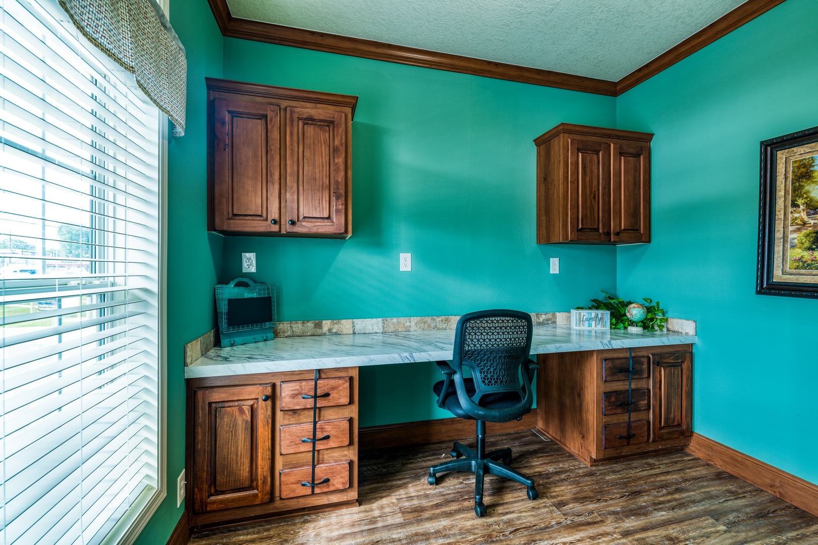 A home office with teal walls features wooden cabinets and a marble countertop. A black office chair and a window with blinds add a cozy, productive atmosphere.