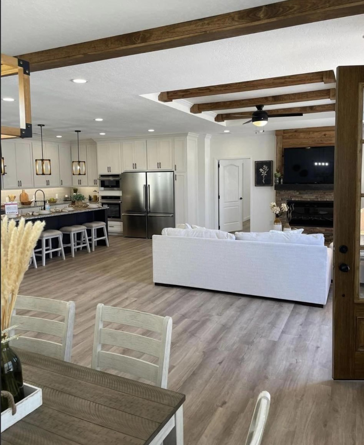 Spacious open-plan kitchen and living area with light wood flooring, exposed wood beams, and white cabinetry. A cozy, modern ambiance is highlighted by a white sofa, large stainless steel fridge, and dark kitchen island with stools.