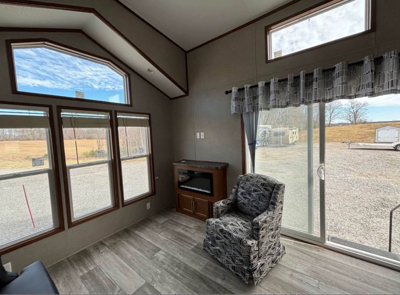 Modern interior with large windows showing a scenic outdoor view. Features patterned armchair, electric fireplace, and gray wood flooring, evoking a cozy ambiance.