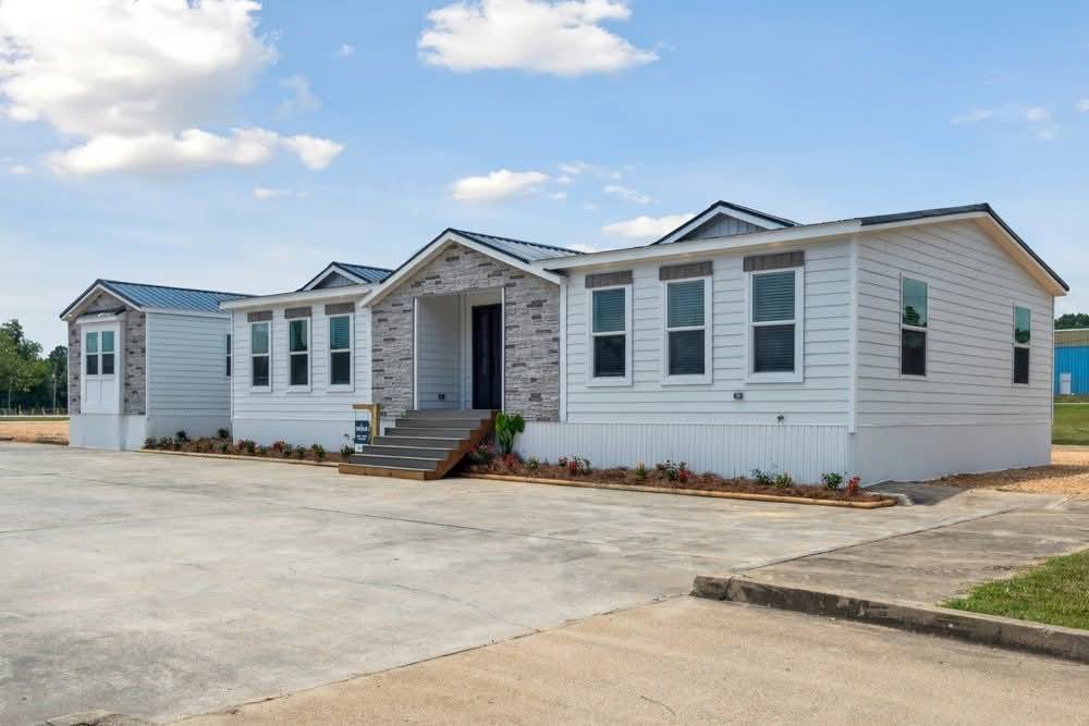 Single-story modular home with white siding, gray stone facade, and a metal roof. It has large windows and a small entrance porch, set on a concrete driveway.