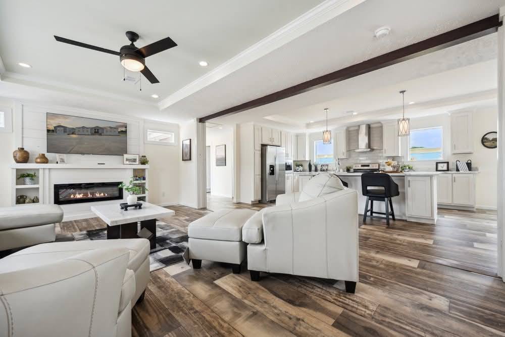 Spacious modern living area with white sofas, a fireplace, and wood flooring. Open kitchen features stainless steel appliances and hanging lights. Cozy yet elegant atmosphere.