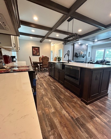 Spacious kitchen with wooden floor, coffered ceiling, and a large island featuring a built-in microwave and sink. Adjacent dining area and cozy living room visible.