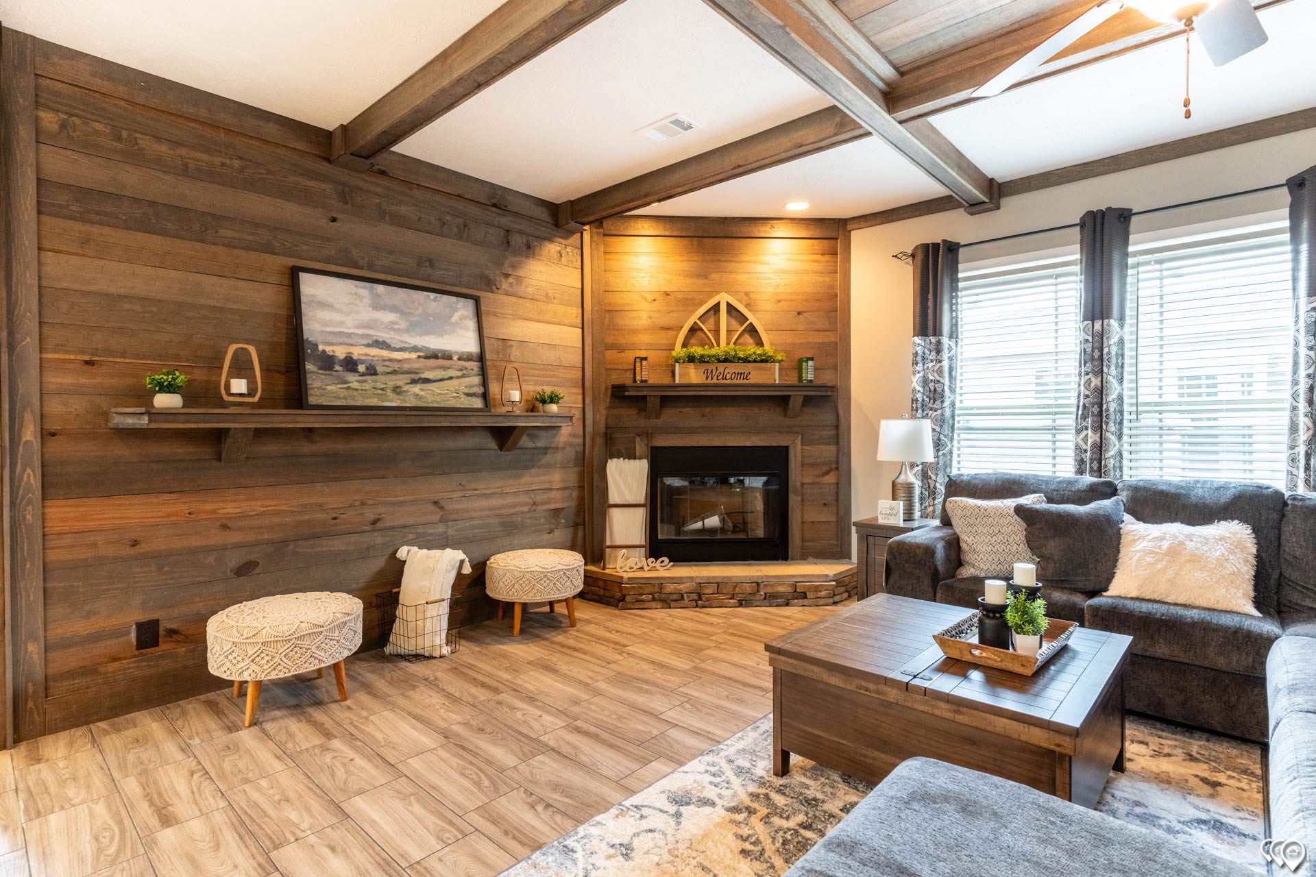 Cozy living room with a rustic wood accent wall, stone fireplace, and beamed ceiling. Plush gray sofas, wooden coffee table, and soft lighting create a warm, inviting atmosphere.