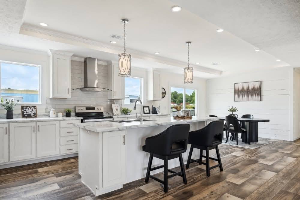 Bright modern kitchen with white cabinets, a marble island, black chairs, and hanging lights. A dining table is in the background. Wood floors add warmth.