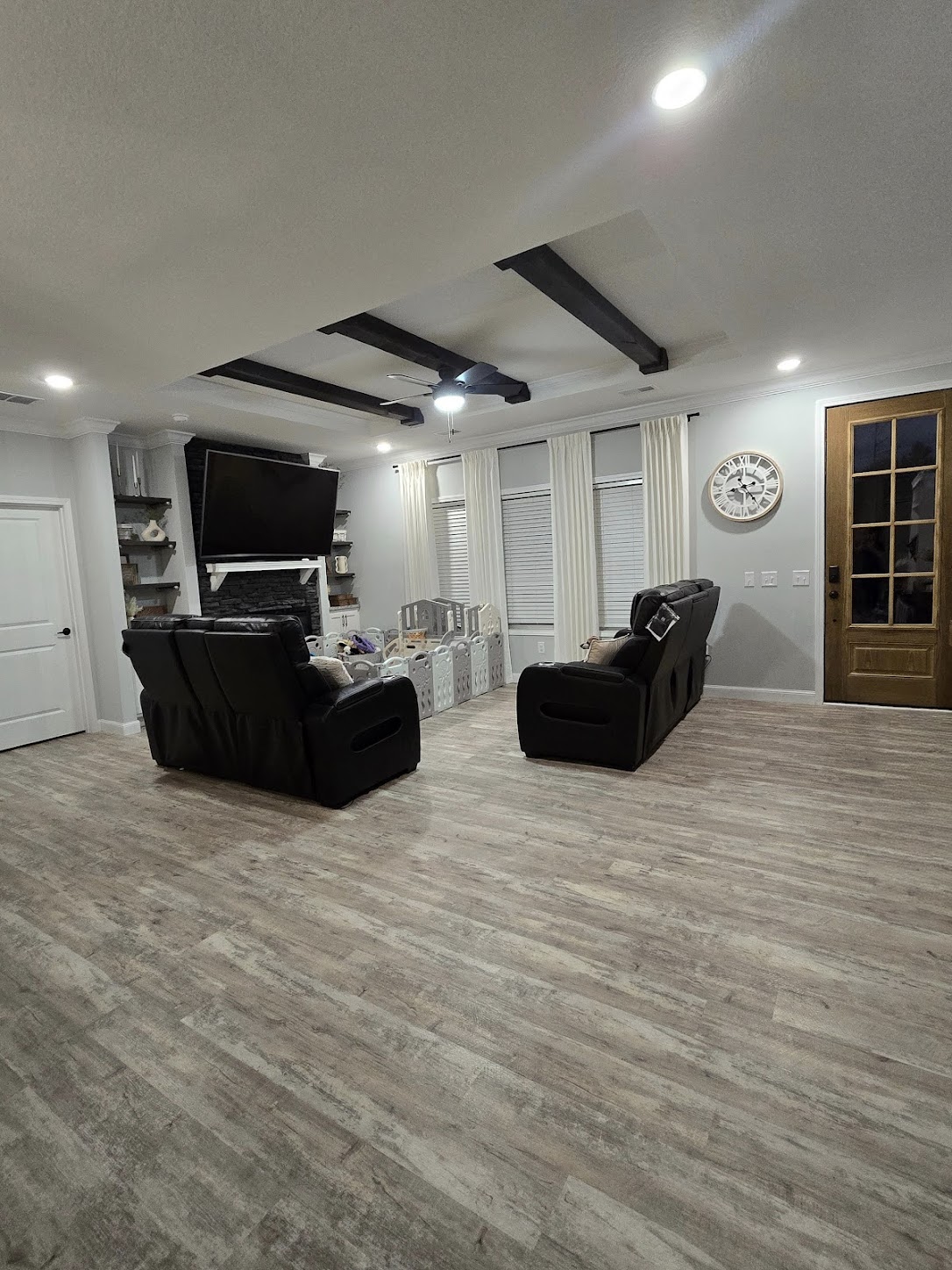 A cozy living room with wooden floors, two black recliners facing a wall-mounted TV, and a rustic clock on the wall. Bright and inviting atmosphere.