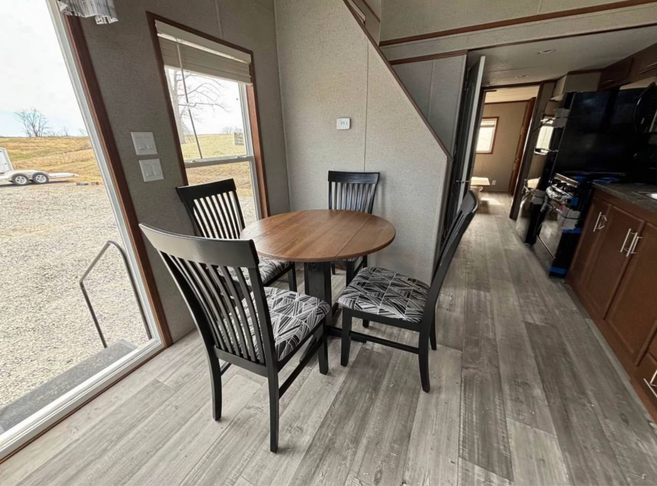Compact dining area with a round wooden table and four cushioned chairs. Adjacent to a large window, light wood floors, and a staircase leading upstairs.