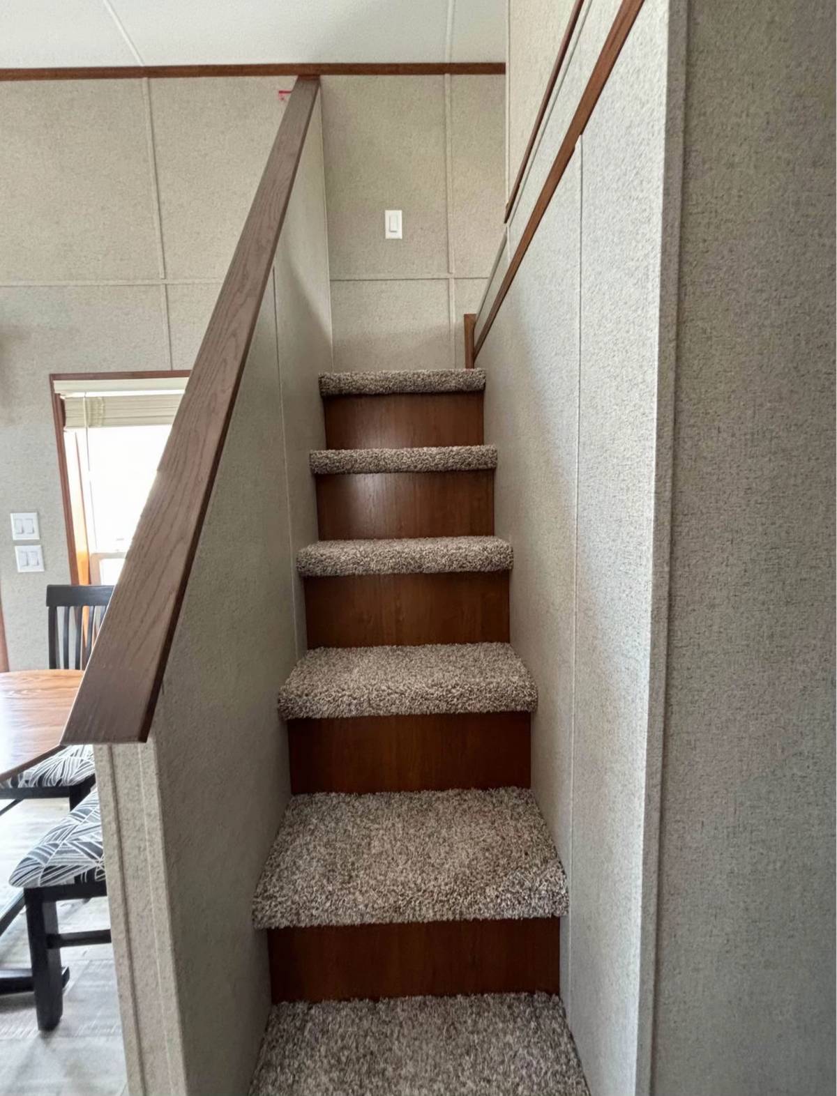 Carpeted brown wooden stairs in a narrow, modern interior. Light walls and ceiling offer a clean, minimalist look. Doorway and chairs are partially visible below.