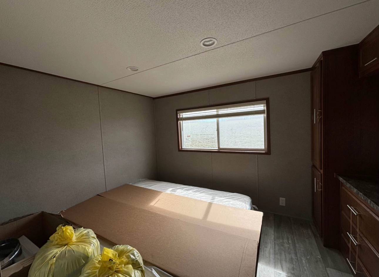 Simple room with a cardboard box and bags on a bare mattress, next to a window with closed blinds. Wooden cabinets stand in the corner.