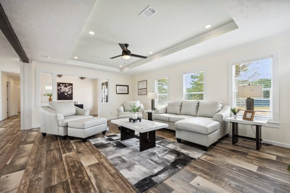 Modern living room with gray sectional sofa, white armchair, and a marble coffee table on a patterned rug. Wooden floor and large windows create a bright, airy space.