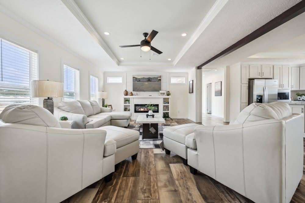 Spacious living room with white leather sofas arranged around a fireplace. Wooden floor, ceiling fan, and abundant natural light create a cozy, modern atmosphere.