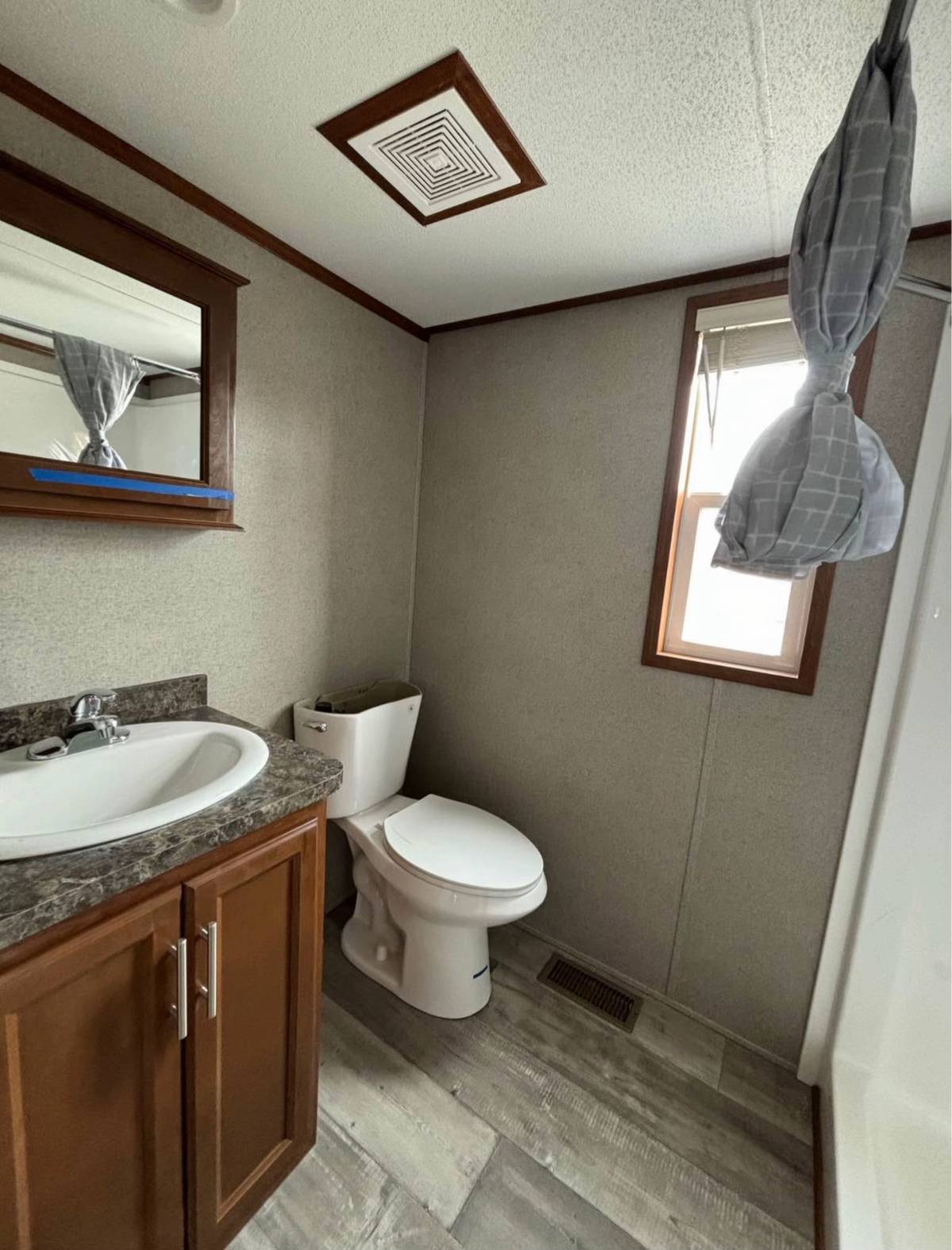 Small bathroom with gray walls, features a toilet beside a wooden vanity with a sink and mirror. Sunlight streams through a window with a tied curtain.