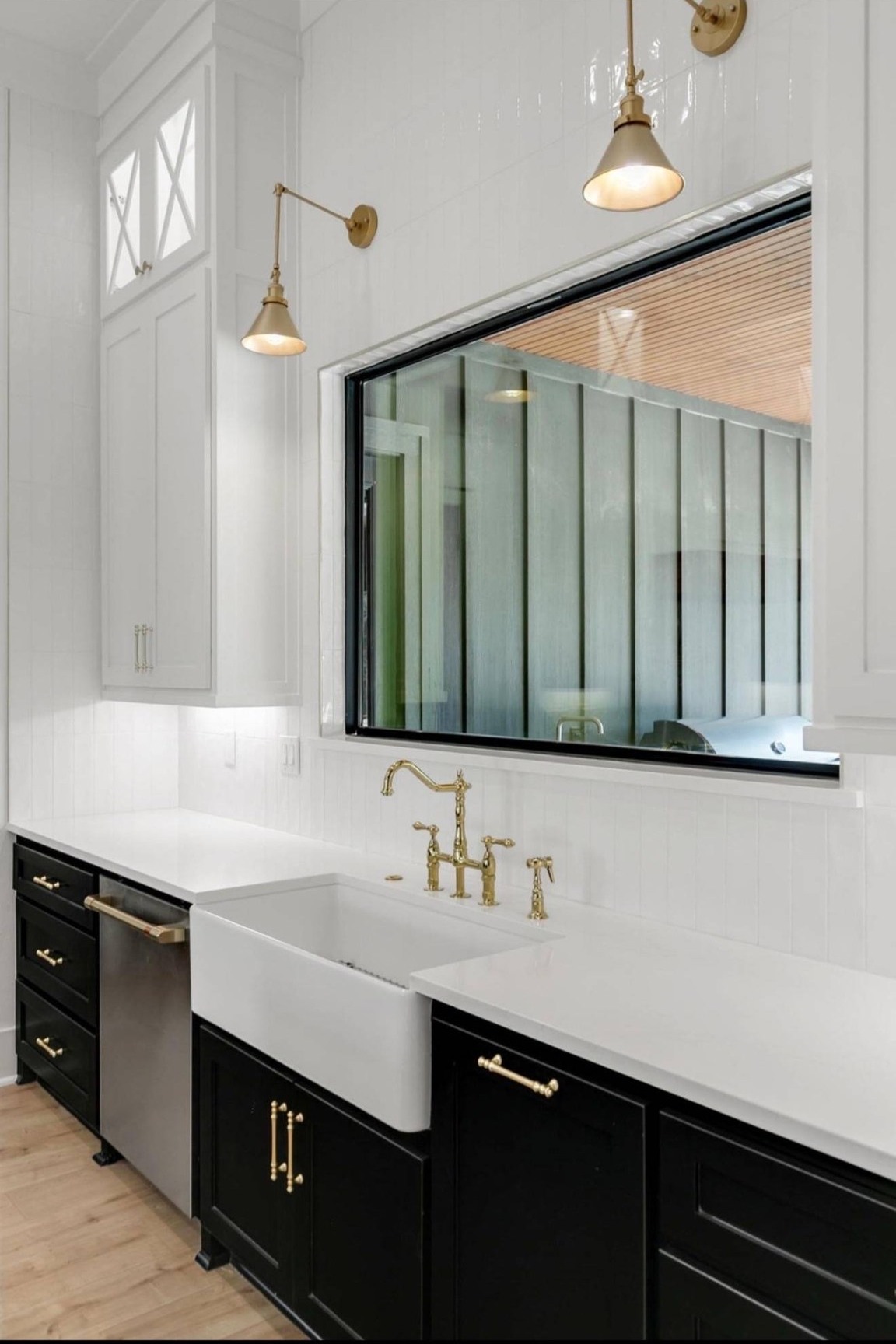 Elegant kitchen with white countertops, black cabinets, and brass fixtures. A farmhouse sink under a large window. Warm lighting from two wall lamps.