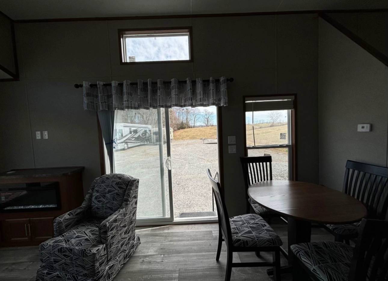 A cozy living room with a patterned armchair, round dining table, and chairs. Large window reveals a sunny outdoor scene with grass and a gravel path.