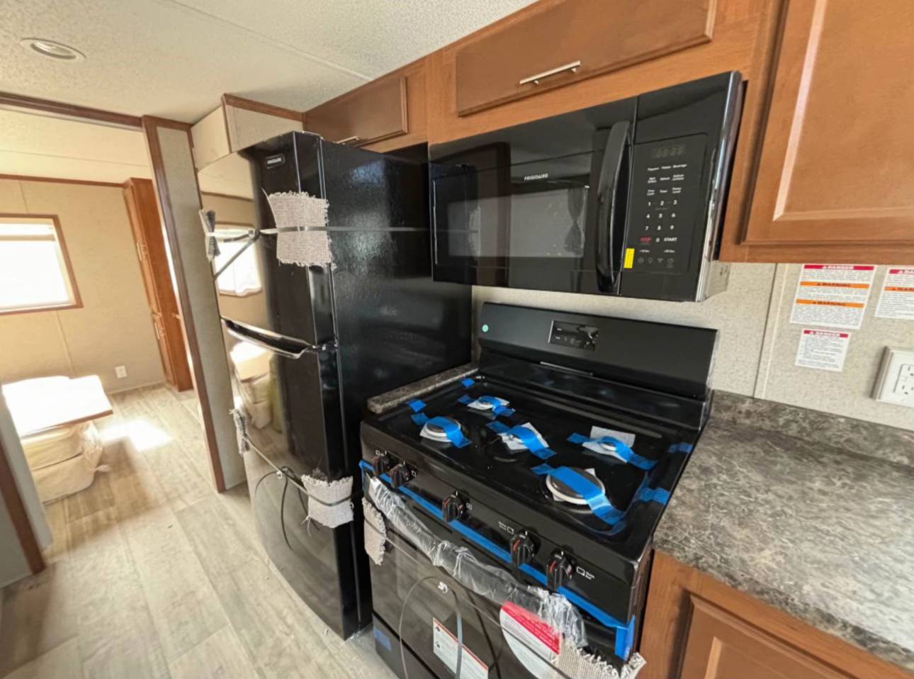 Modern RV kitchen featuring a black fridge, stove, and microwave, all with protective tape. Light wood flooring and natural light create a cozy feel.