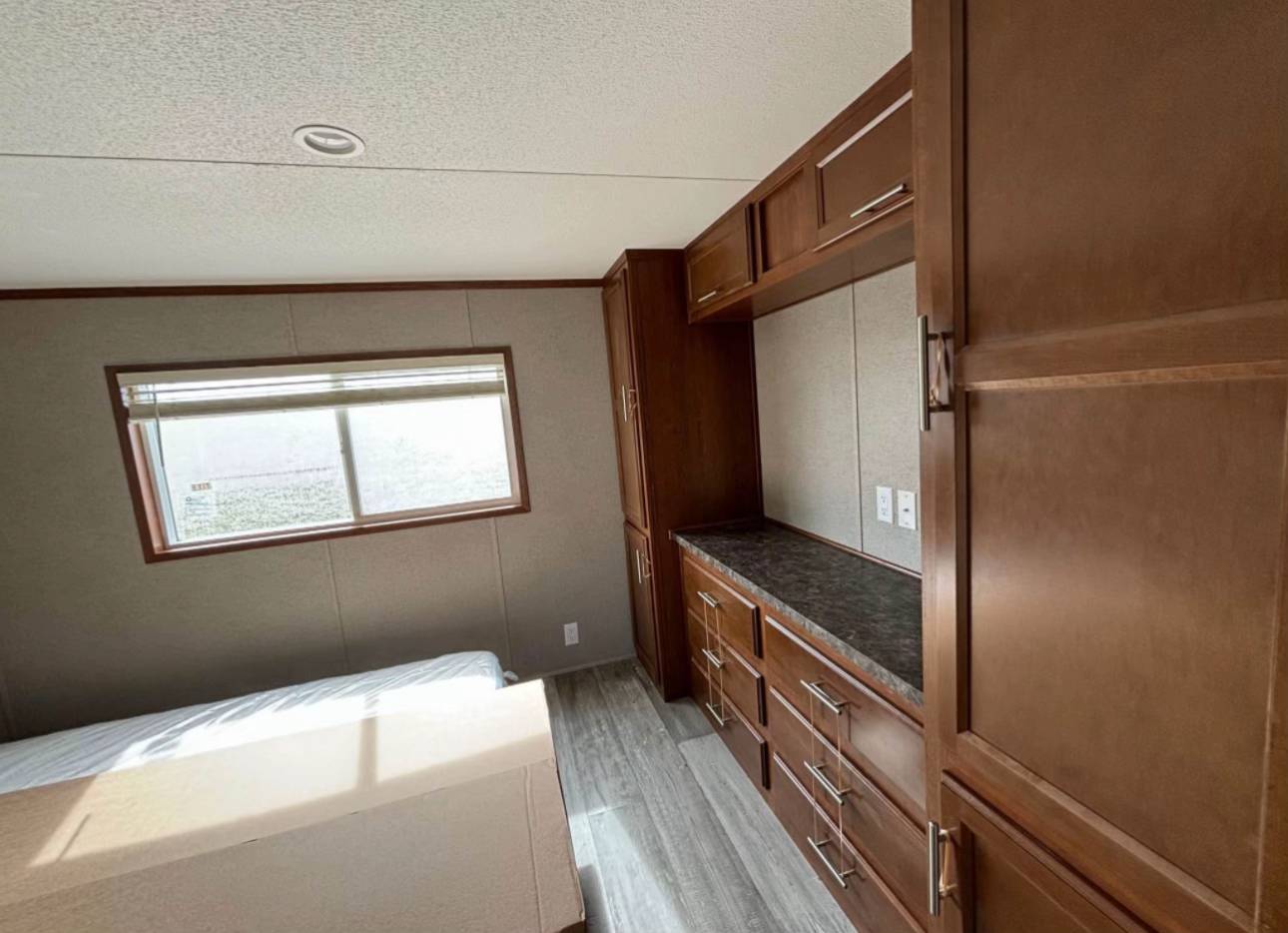 Bedroom with brown wooden cabinets and drawers on the right, a bed on the left, gray flooring, and a window letting in natural light, creating a cozy tone.