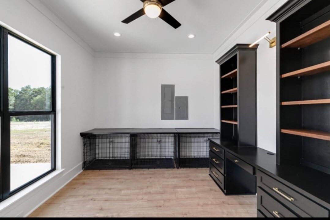 A minimalist office space with a dark wood desk, black paneled wall with cabinets, light wood flooring, and two windows showing a green landscape.