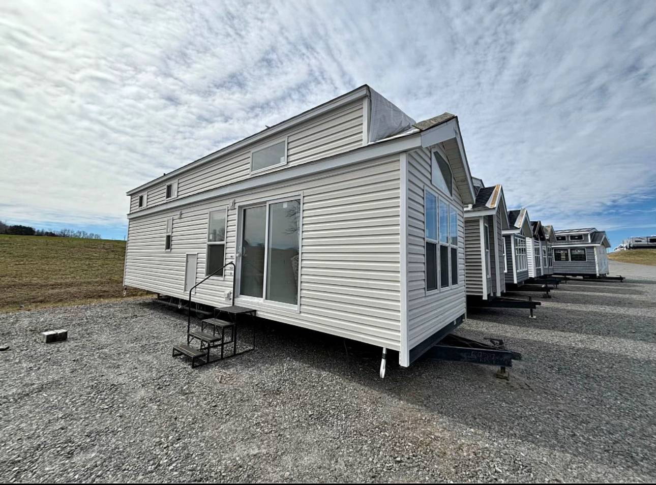 Row of white, compact tiny houses with large windows, lined up on a gravel path under a cloudy sky. The setting conveys a sense of simplicity and community.