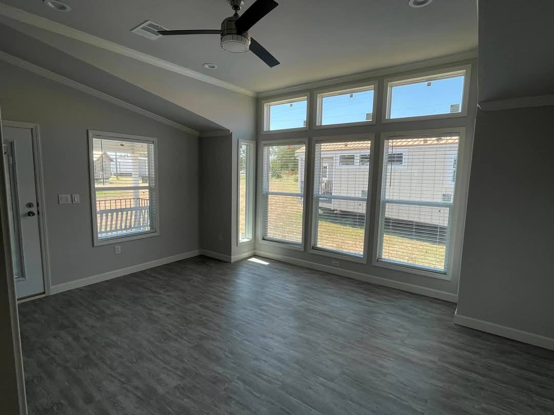 Bright room with large windows, gray wood floor, and ceiling fan. Natural light floods the space, creating a calm and open atmosphere.