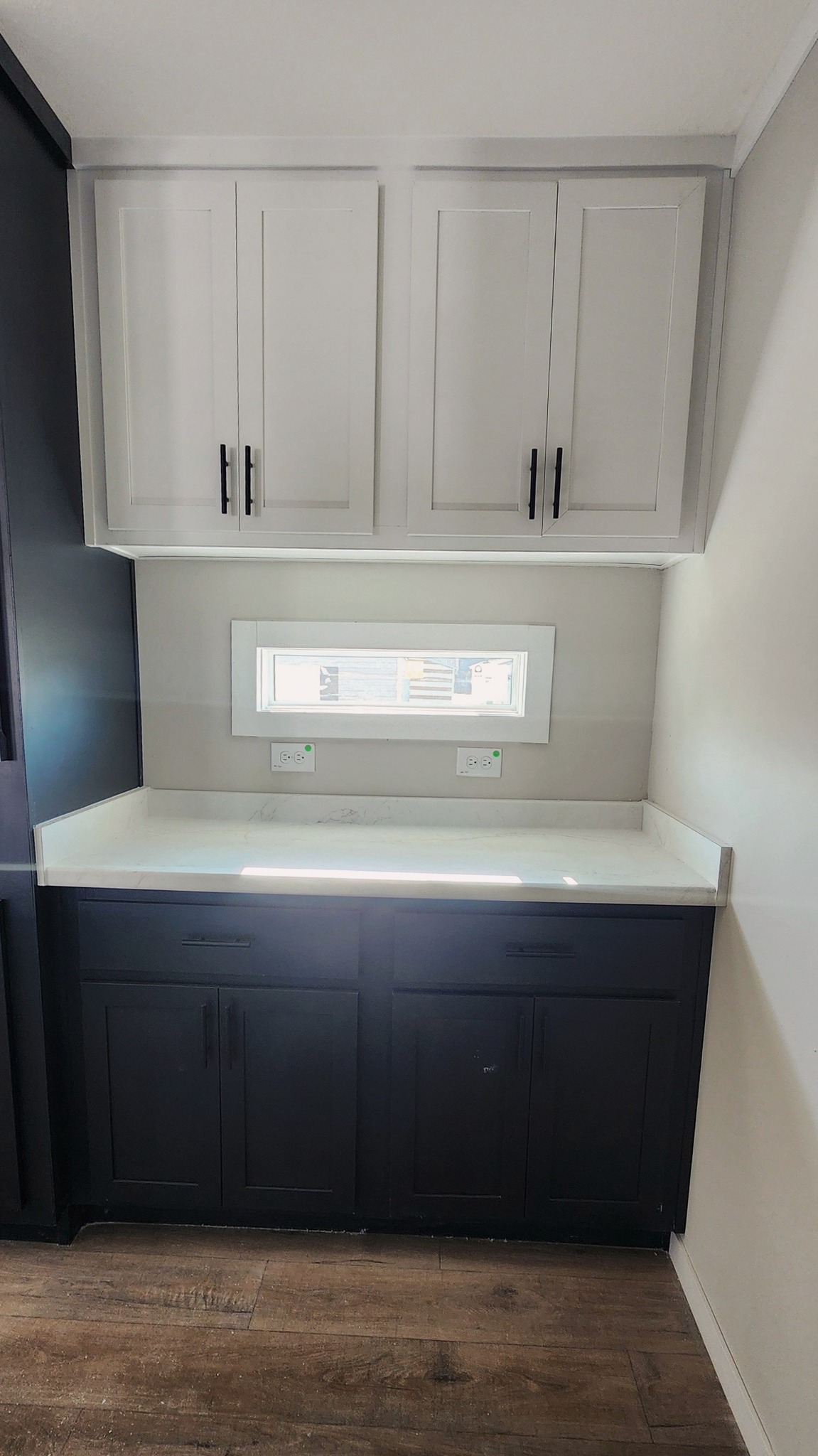 Compact kitchenette with white upper cabinets, dark lower cabinets, and a small counter. A narrow window above the counter lets in natural light.