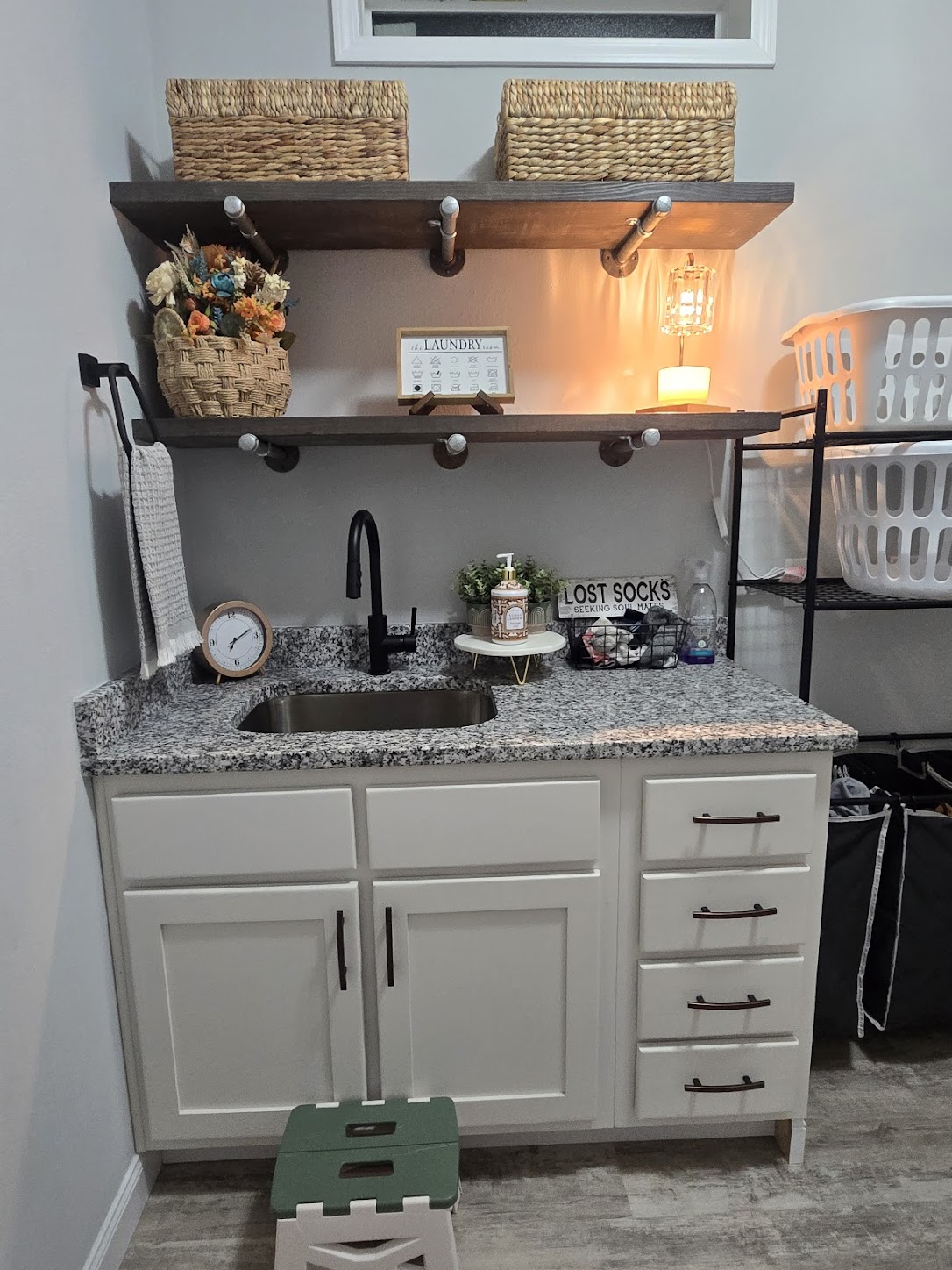 Compact laundry room with granite countertop, white cabinets, black faucet, and wall shelves. Warm lighting, woven baskets, and a "Lost Socks" sign add cozy charm.