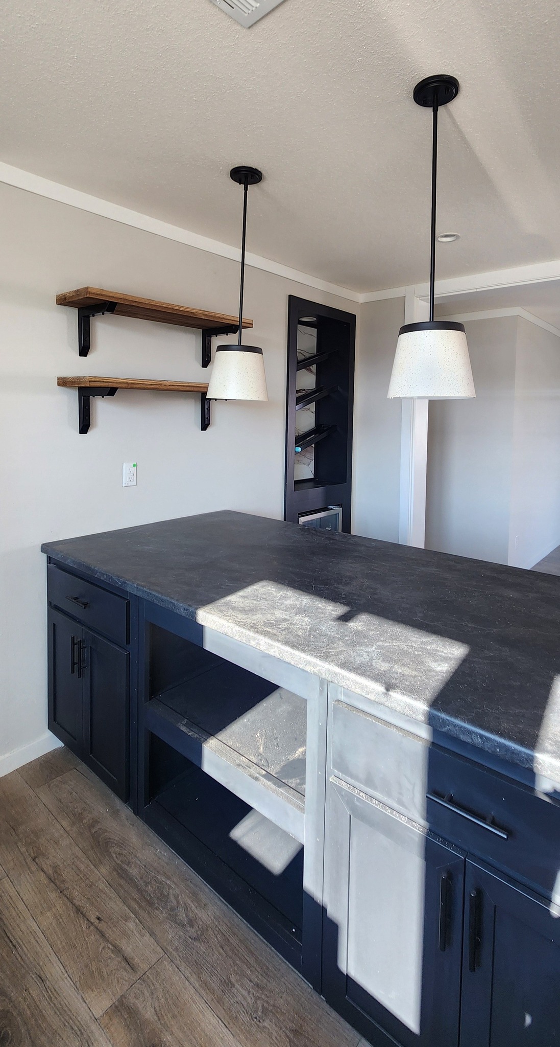 Modern kitchen with dark blue cabinets, a sleek gray countertop, and two white pendant lights. Wooden shelves and natural wood flooring add warmth.