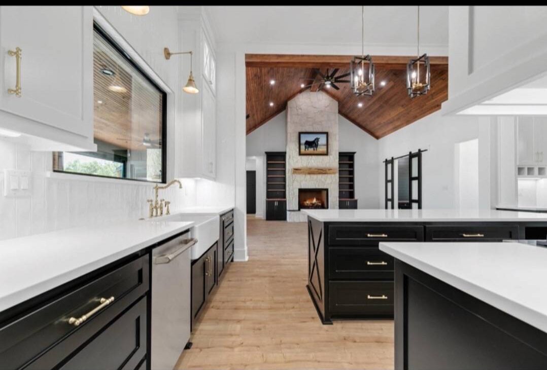 Sleek kitchen with black and white cabinetry, gold fixtures, wood flooring, and modern pendant lighting, leading to a cozy living room with a fireplace.