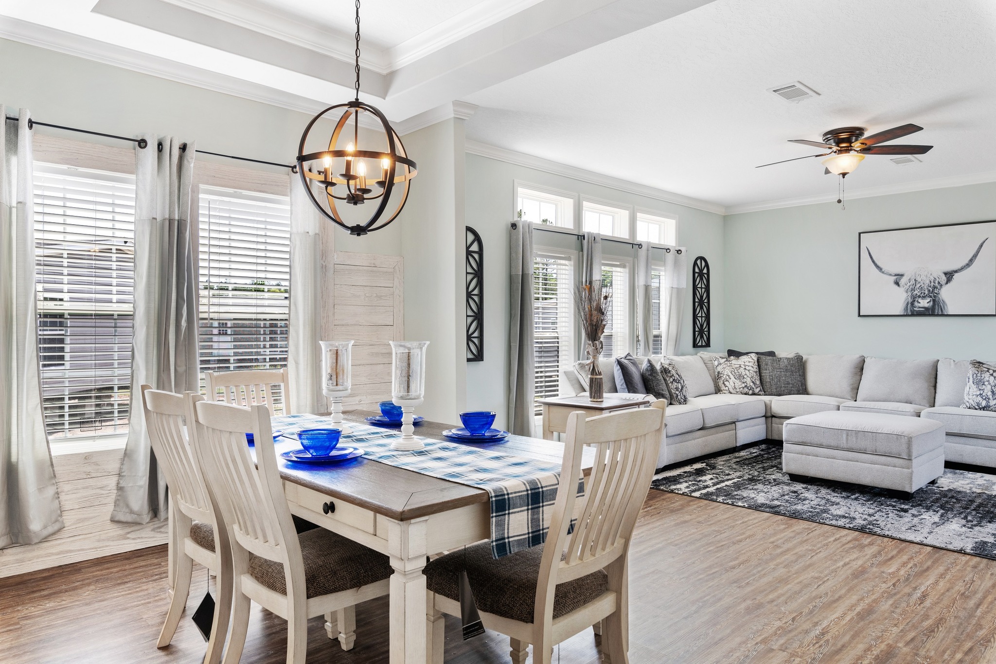Bright living-dining area with a rustic table set, blue accents, and a plaid runner. Cozy gray sectional, cow artwork, wooden floors, and modern chandelier.