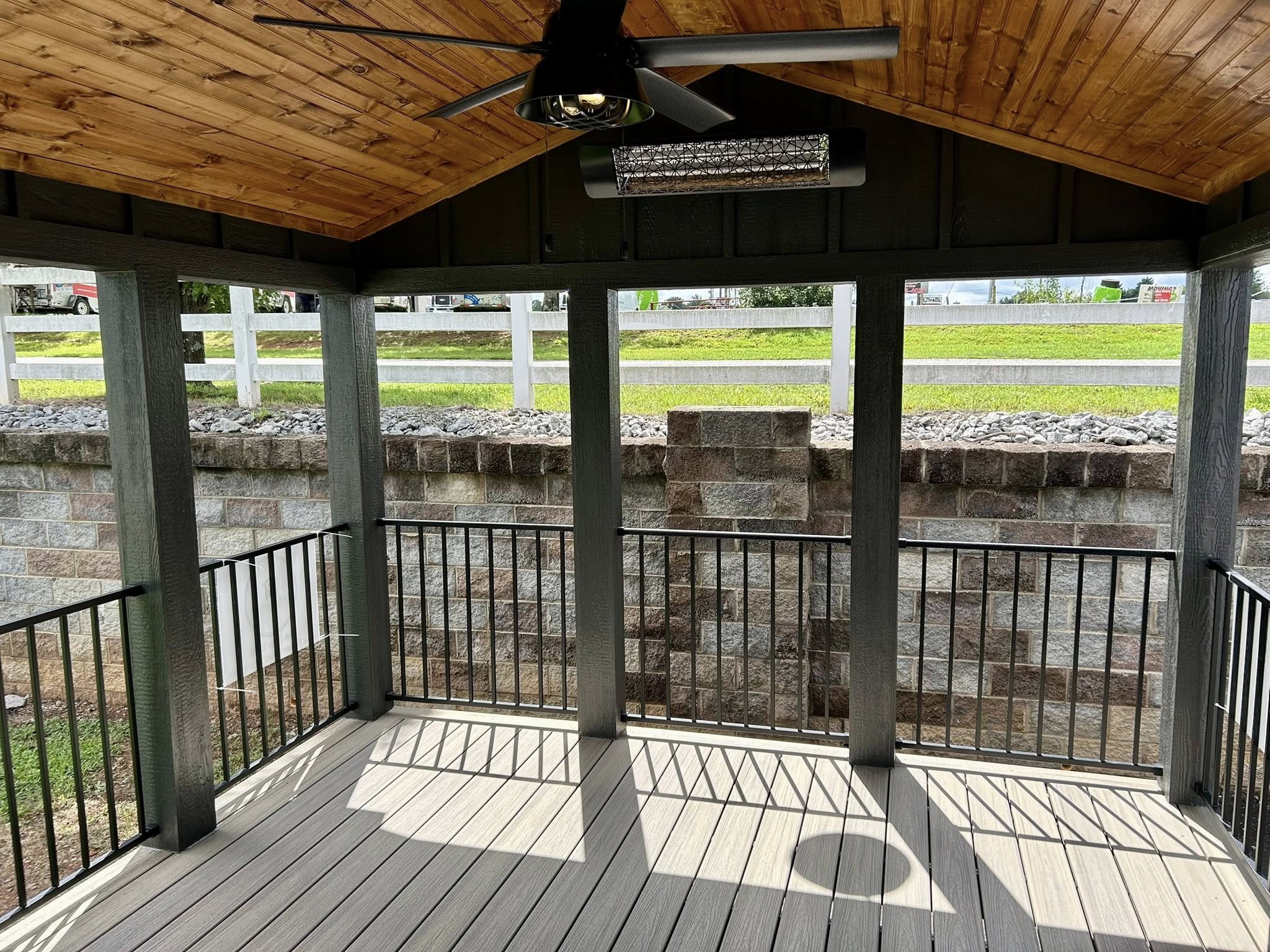 Cozy gazebo with a wooden ceiling, fan, and heater. Black railing surrounds the gray deck. Sunlight casts shadows on the floor, creating a warm, inviting feel.