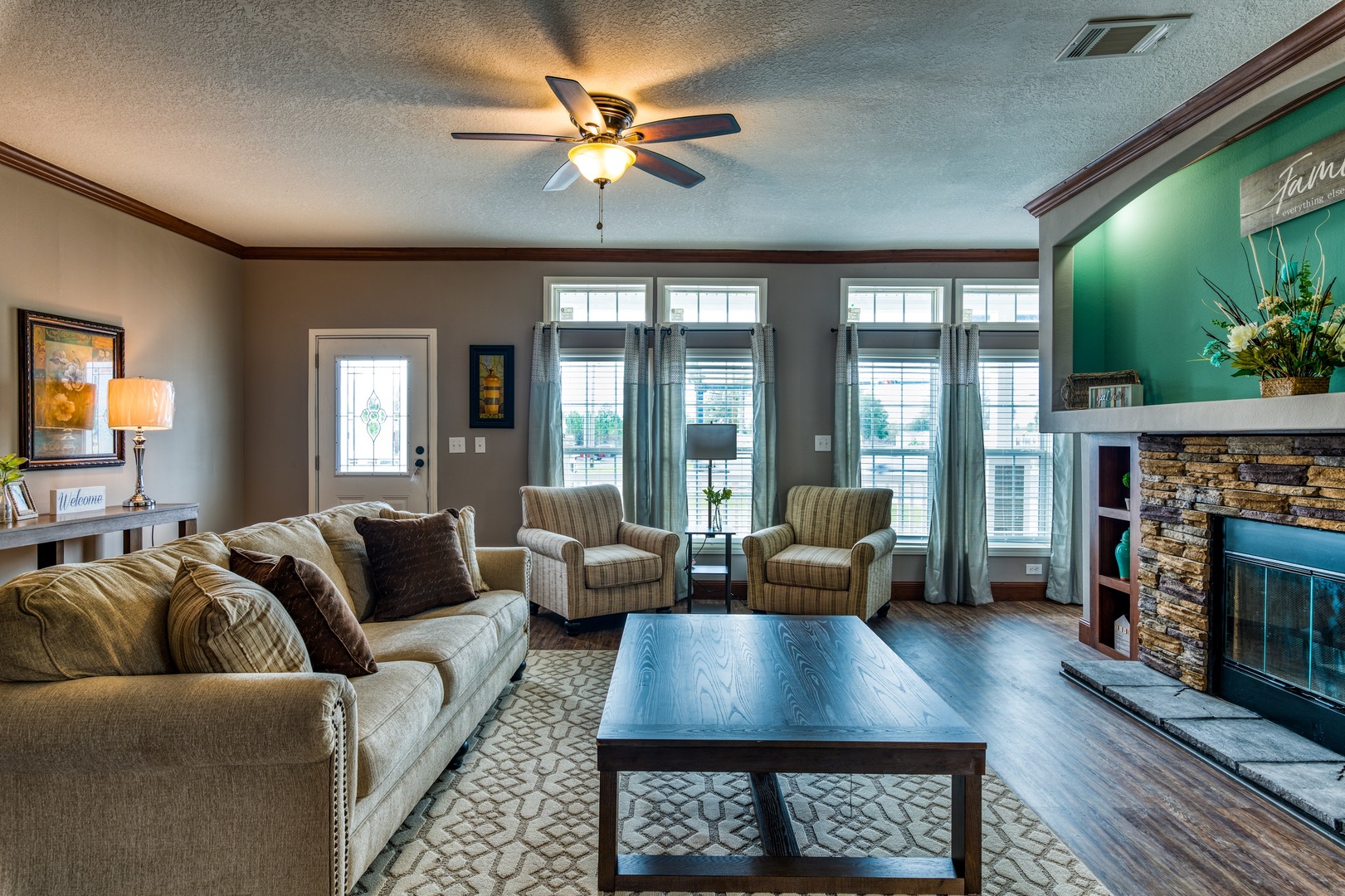 Cozy living room with a beige sofa, two armchairs, and a wooden coffee table. A stone fireplace and large windows bring warmth and natural light.