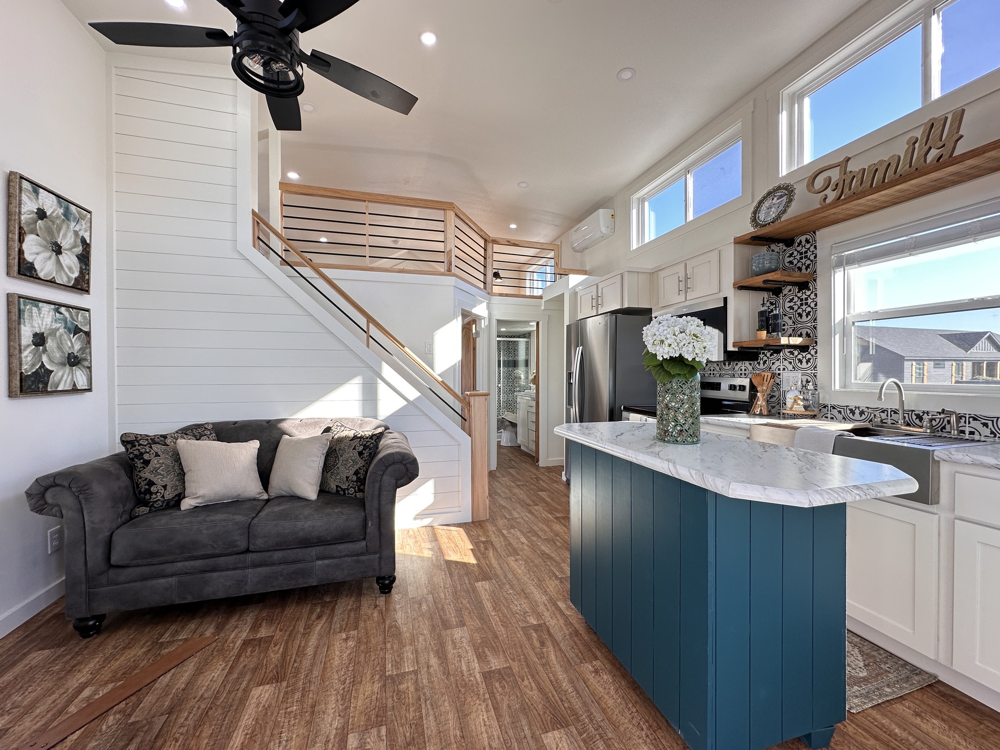 Compact, modern tiny home interior with a cozy gray sofa, wooden stairs to a loft, turquoise kitchen island, and bright windows. Welcoming atmosphere.