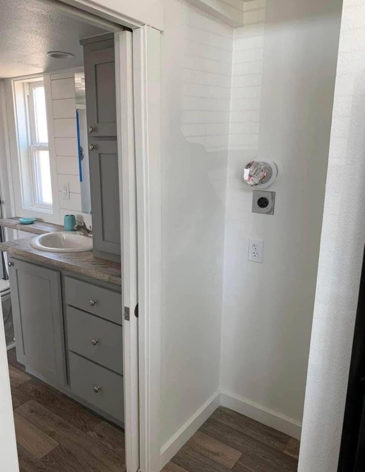 Compact bathroom with gray cabinetry, a sink, and wooden flooring. A small corner features two round wall vent covers, one with a playful paper cover.