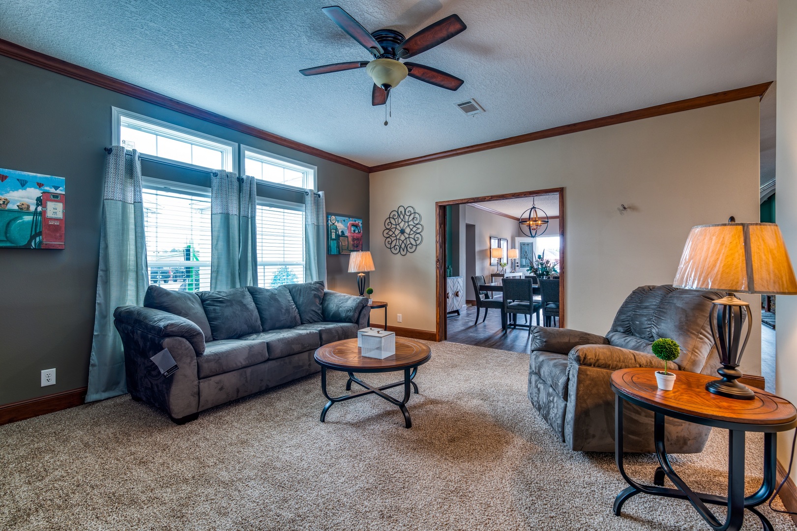Cozy living room with a plush sofa and recliner, wooden coffee and side tables, soft lighting from lamps, and a ceiling fan. Dining area visible through archway.