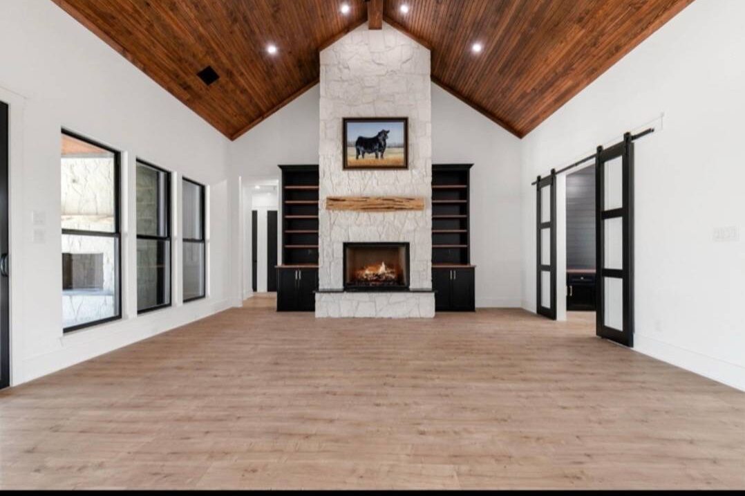 Spacious living room with vaulted wooden ceiling, stone fireplace, and mounted TV. Large windows on the left and glass doors on both sides convey openness.