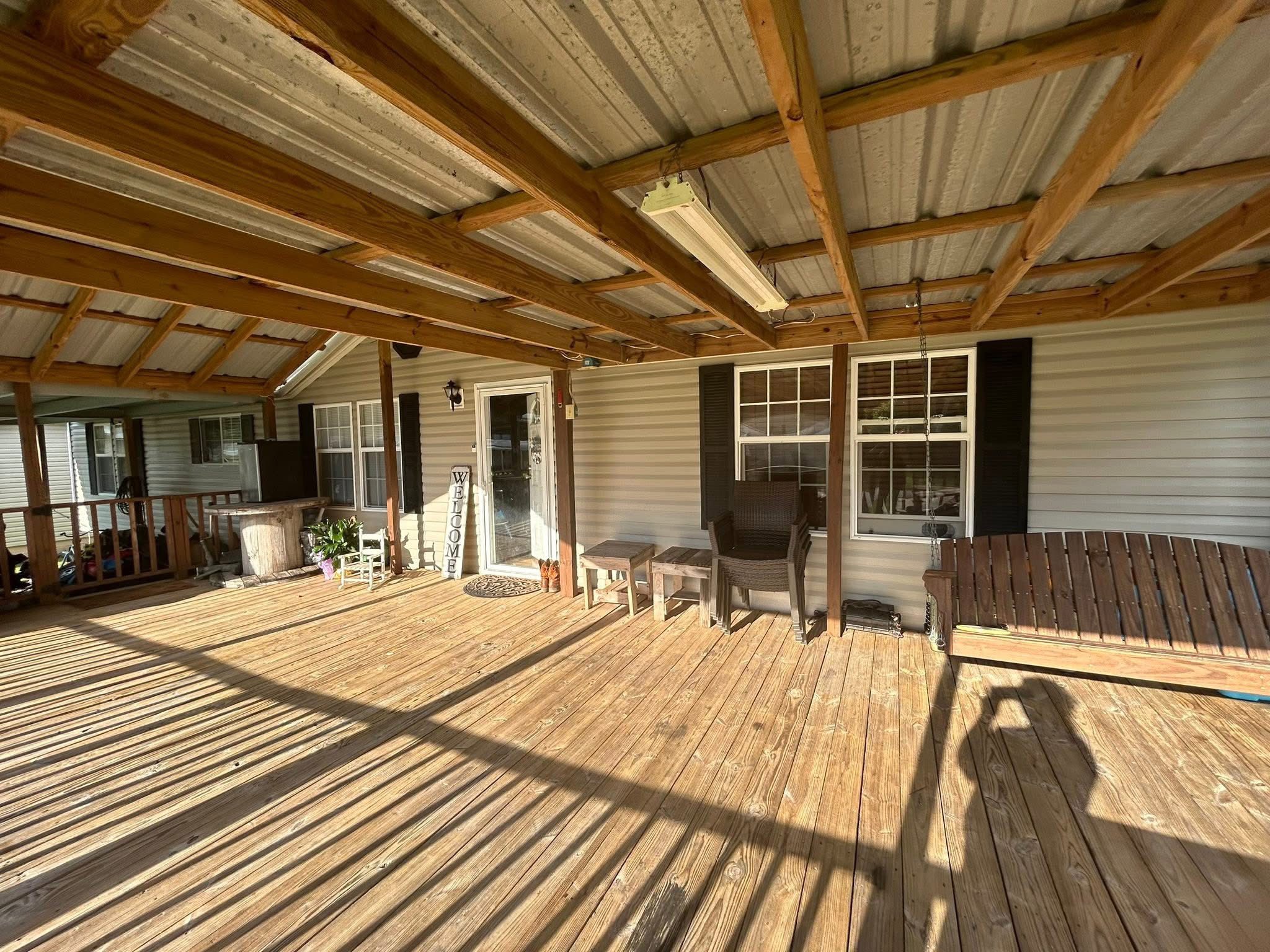 Covered wooden porch with sunlight casting long shadows. Features wicker furniture, a wooden swing, potted plants, and a welcome sign by the door.
