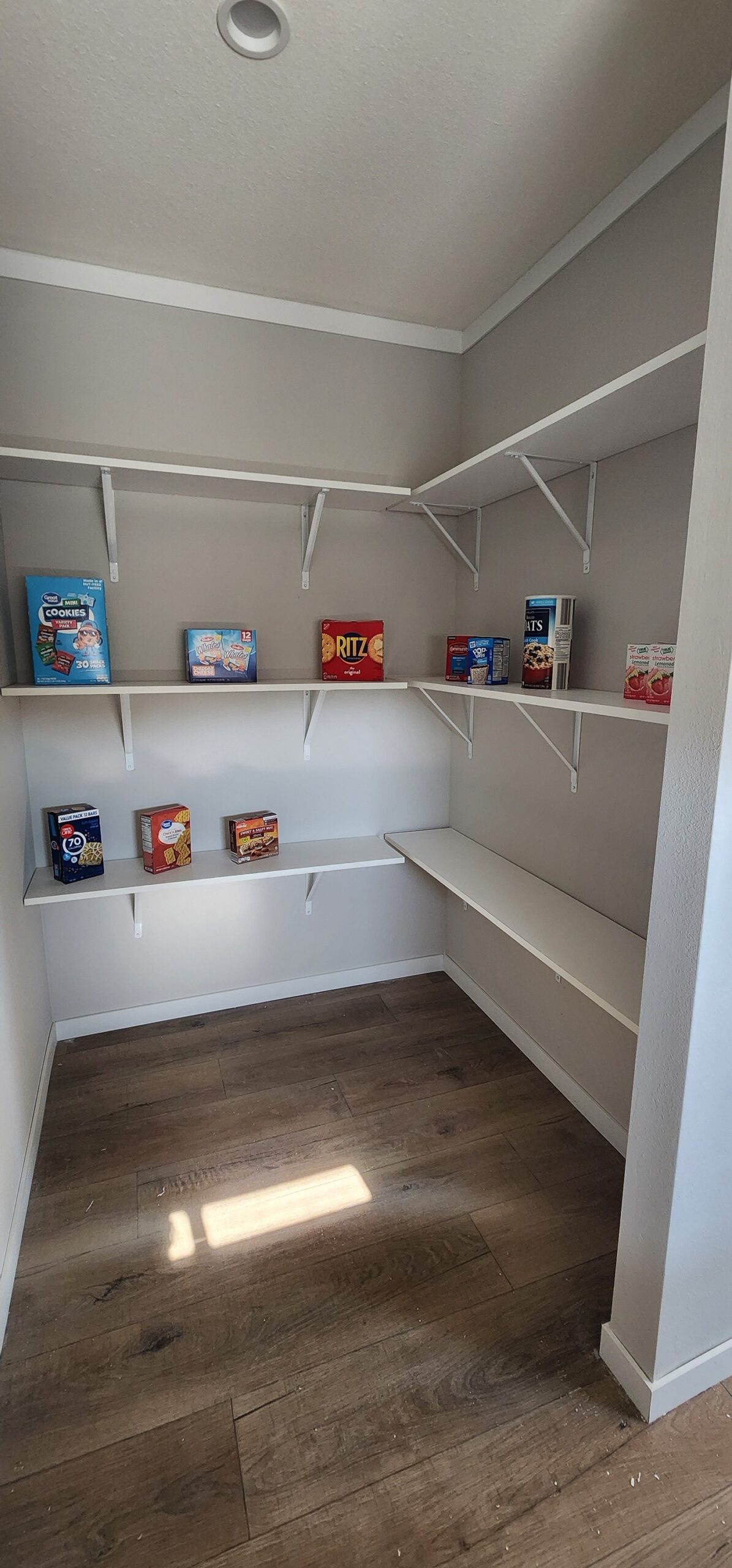 A pantry with wooden floors and white walls has three shelves. Sparse items include cereal boxes, crackers, and oatmeal, creating an organized yet minimal feel.