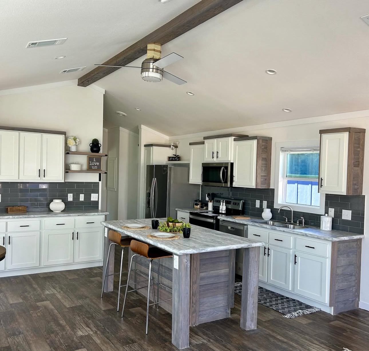 Modern kitchen with white and wood cabinets, gray backsplash, and a central island. Features stainless steel appliances, bar stools, and a ceiling fan.