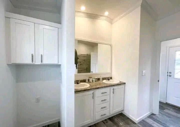Bright bathroom with dual sinks, large mirror, white cabinetry, and wood floor. Soft lighting from recessed ceiling lights creates a clean, modern feel.